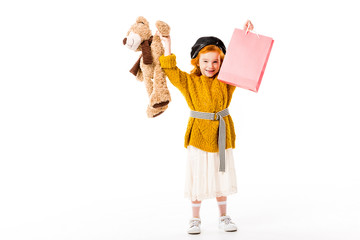 happy redhead child holding shopping bag and toy in hands above head isolated on white