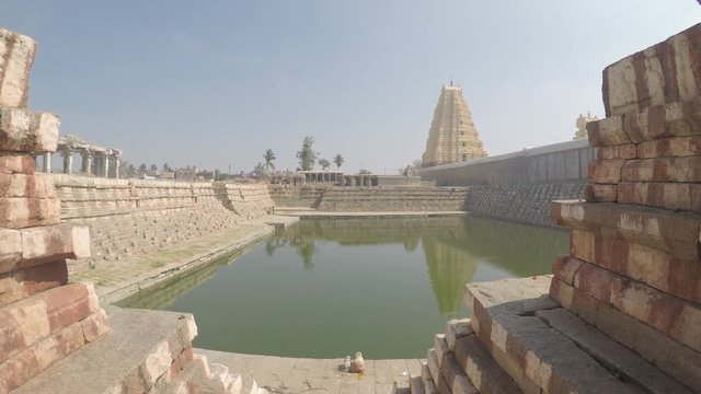 Sacred water tank with Virupaksha temple in background, smooth tracking shot from high up descending to water level, in Hampi, India