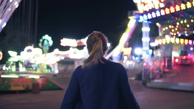 Back view of young attractive girl with ponytail walking forward then turning around smiling and looking at camera hanging out in amusement park with attractions background. Slowmotion shot