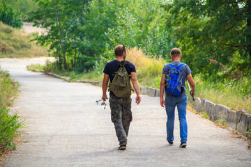 Fototapeta premium Two fishermen go fishing with fishing rods along the asphalt path through the forest.