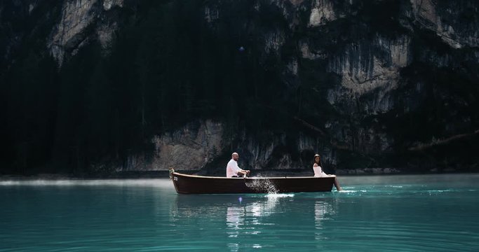 Romantic attractive couple in the middle of the lake, with a wooden boat the man are rowing and his girlfriend are playing with legs in the water. 4k