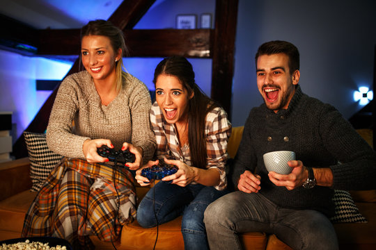 Two female best friends sitting at home on pleasant evening and playing games on console.They challenge each other to win while man cheering.