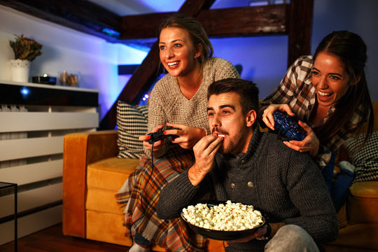 Two female best friends sitting at home on pleasant evening and playing games on console.They challenge each other to win while man cheering and eat popcorn.