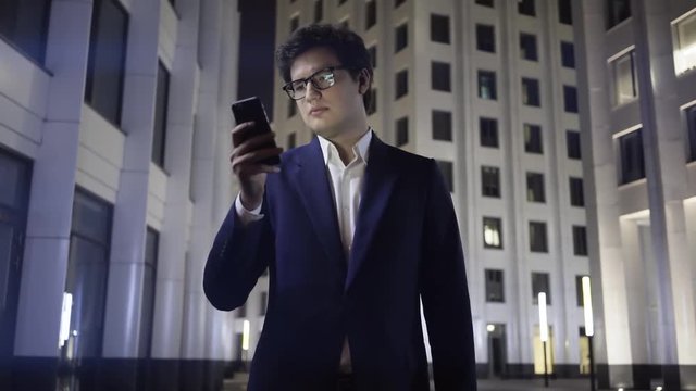 Handsome young man wearing a suit and glasses is looking at his smartphone screen and frowning while standing outside at night. Handheld real time establishing shot