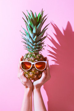 Creative layout made of summer tropical fruits and leaves. Pineapple in glasses on a woman hand. pink isolated bacground. Flat lay. Food concept.