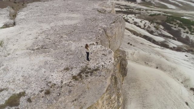 Young woman with long hairs is standing at the edge of white rock with raised hands and enjoying the scenery. Aerial view. Drone is flying over precipice with camera tilt.
