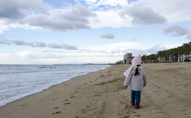 Little girl wear jacket and scarf walk on sand beach near mediteranean sea with waves on cloudy sky in winter time