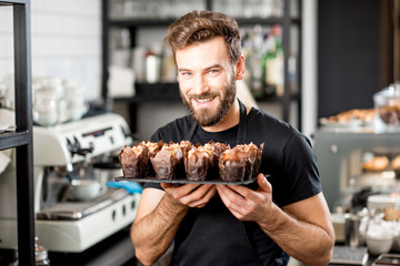 Portrait of a handsome bearded confectioner in black t-shirt and apron holding mafins at the cafe