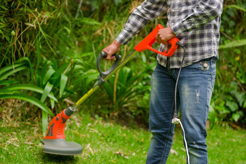 Obraz premium Young worker wearing jeans and long sleeve shirt and using a lawn trimmer mower cutting grass in a blurred nature background