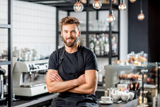 Portrait of a handsome barista in black t-shirt and apron sitting at the bar of the modern cafe