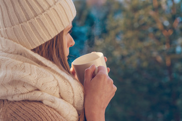 Young woman wearing warm knit clothes drinking cup of hot coffee outdoors in sunlight.
