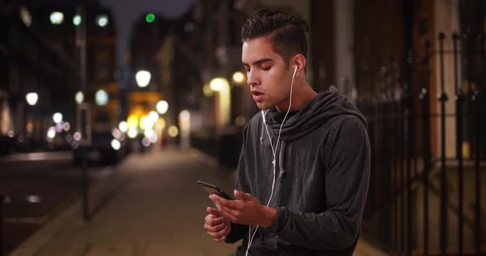Millennial Latino jogger on city street at night listening to music on cellphone