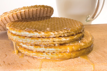 round cookies or wafers with a layer of caramel lies on a rough pile on a wooden board, mug of clear glass with milk