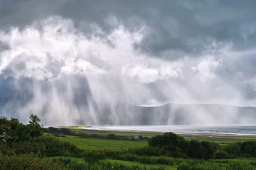 Distance view of a rain storm. Pouring down water over mountains, sun rays, beach. Ring of Kerry, Ireland