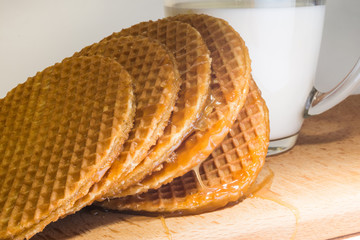 round cookies or wafers with a layer of caramel lies on a rough pile on a wooden board, mug of clear glass with milk
