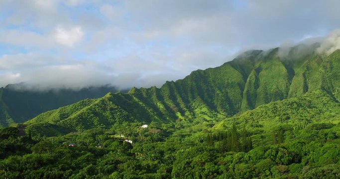Aerial shot of vibrant green Koolau mountain cliffs bathed in early morning golden hour light, with white mountain fog forming on the high peaks, against a partly cloudy blue morning sky. Kaneohe, Hi.