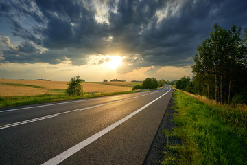 Empty asphalt road leading around golden cornfields in rural landscape at sunset with dramatic...