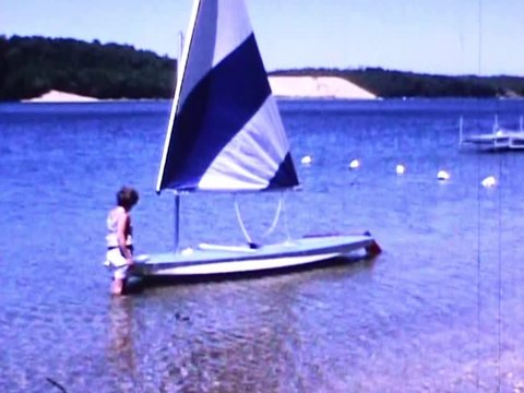 A woman stands in freshwater pond by the shoreline holding sunfish sailboat waiting to go sailing on summer day in 1971