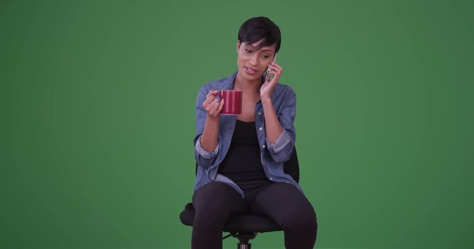 Millennial black woman smiles while having a conversation on her mobile phone and drinks coffee on green screen. On green screen to be keyed or composited. 