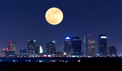 Night view of the Tampa Florida skyline showing skyscrapers with lights and a huge full moon in the sky over the buildings. - 184878139