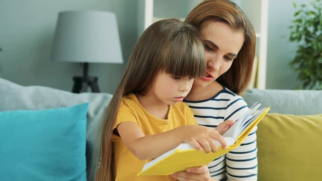 Little pretty girl reading a book together with her mother while sitting on her knees in the living room. Portrait shot. Indoors. Close up