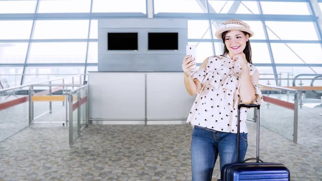 Happy young woman taking selfie photo in the airport terminal with her suitcase and wearing a hat