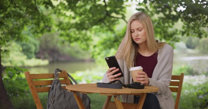 Pretty Caucasian girl drinks coffee and browses web on phone in public park