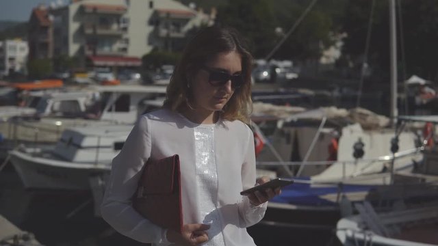 positive business woman with smartphone and handbag in formal clothes standing on the background of blurry boats