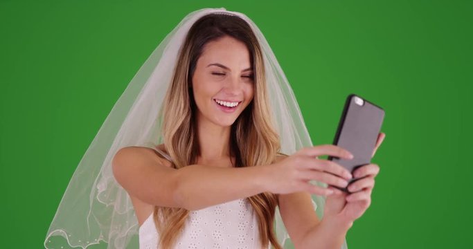 Bride in wedding dress and veil taking a selfie with cell phone on green screen. On green screen to be keyed or composited.