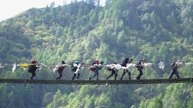 Sherpas and hikers walk a bridge crossing a valley in the foothills of the Himalayas. They travel in mountains and carry big backpacks