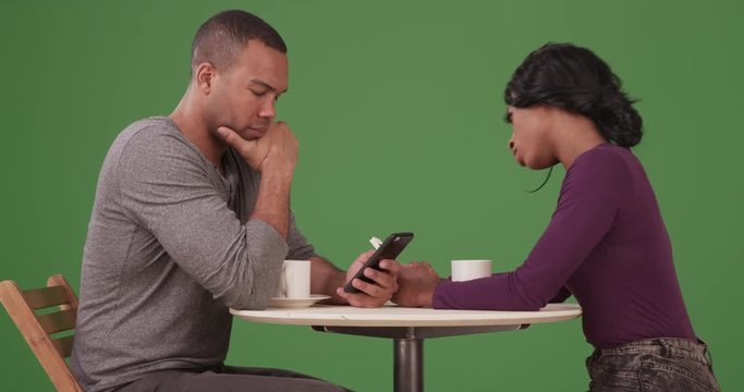 Black couple drinking coffee and browsing internet on smartphones on date on green screen. On green screen to be keyed or composited. 