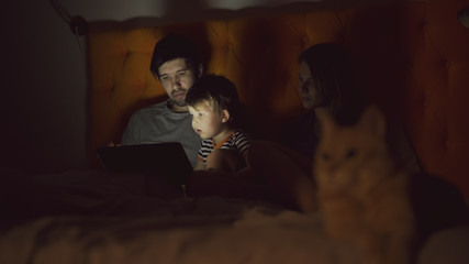 Happy family with little son learning to play tablet computer lying in bed with cat at home
