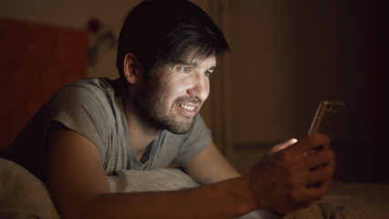 Closeup of Young smiling man using smartphone for surfing social media lying in bed at home at night
