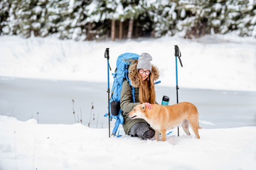 Woman having a break during the winter hiking stroking her dog at the snowy mountains near the lake and forest