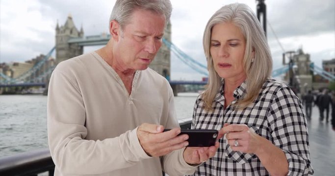 Old white male and wife swipe through photos on phone near Tower Bridge