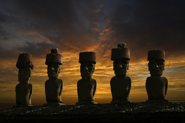 Fünf Moai Statuen mit Kopfbedeckung vor einem dramatischen Wolkenhimmel am Abend.
