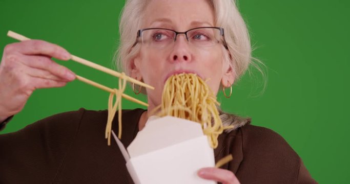 Slow motion of mature woman messily eating noodles on greenscreen. Close up of Caucasian woman in her 50s eating Chinese takeout on green screen to be keyed or composited. 