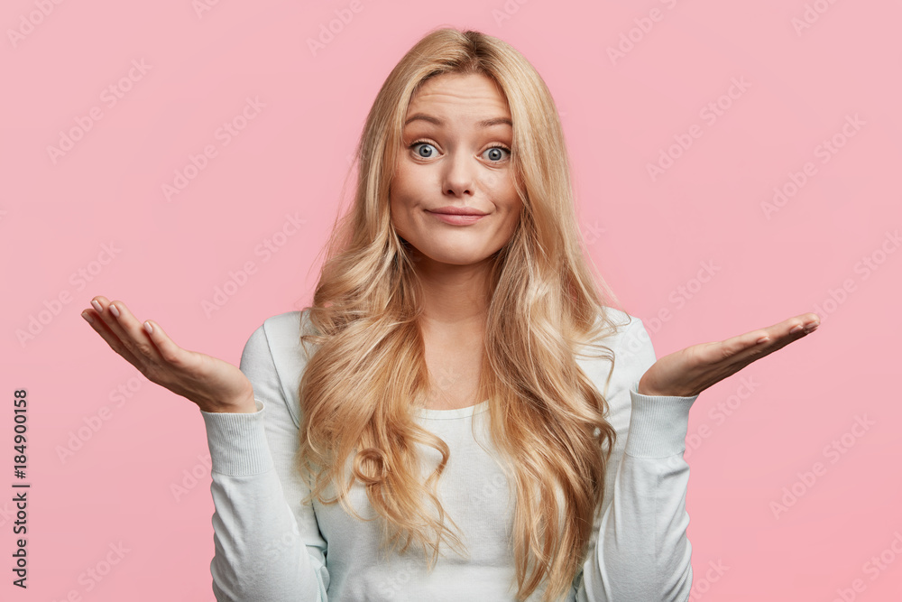 Wall mural Pretty blonde female with blue eyes and blonde hair, shrugs shoulders, being doubtful about making serious decision, feels uncertain, isolated over pink background. Hesitation and confusion. - Wall murals