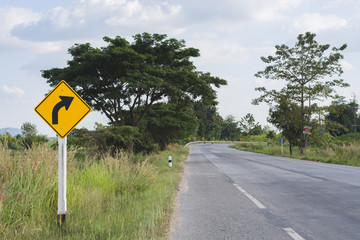 a winding road with a yellow sign indicating a sharp curve to the left. There are trees on either side of the road.