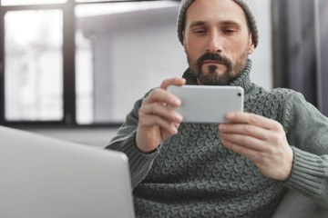 Mature bearded male uses smart phone for making photos, sits in front of laptop computer, uses free wifi, checks camera on mobile. Middle aged unshaven man in stylish clothes indoor, uses technology