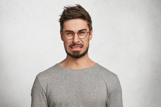 Portrait of handsome bearded male with disgusting expression sees sour lemon in front, frowns in displeasure, isolated over white concrete background. Male expresses aversion towards something