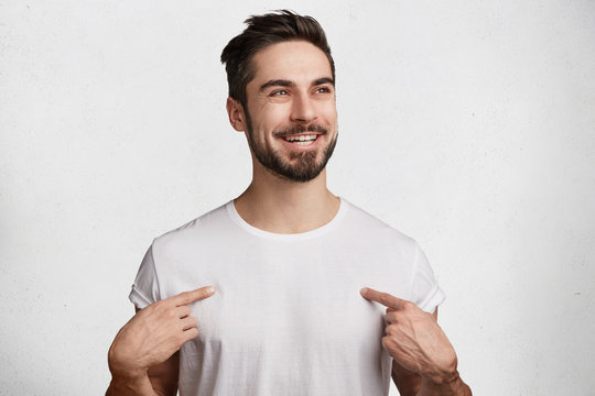 Glad positive male with beard and mustache wears casual white t shirt, has shining happy eyes and pleasant broad smile, indicates at copy space for your design. People, advertisment, positiveness