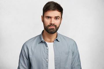 Portrait of serious bearded male wears fashionable denim shirt, hears attentively something important, isolated over white concrete wall. Young male student with thick beard and mustache indoor