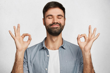 Positive bearded male model wears denim shirt, makes ok gesture and closes eyes, has happy expression, isolated over white background. Optimistic male expresses his approval. Body language concept