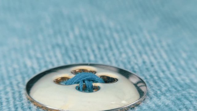 Sewing needle with a blue thread sews a button of gold color with the help of a thimble to a jeans clothes. Macro. Closeup. Shallow depth of field