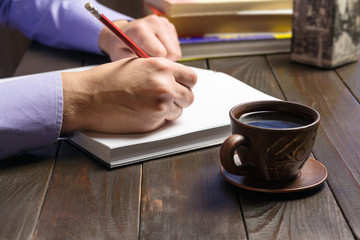 Fototapeta na wymiar Male hand with red pencil writes in notepad or book with white clean pages, black coffee in brown cup on dark wood working table, working or study concept