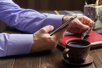Home office workplace, male hands hold a smartphone and credit card, notepad, cup of black coffee on dark wood working table, business, finance concept
