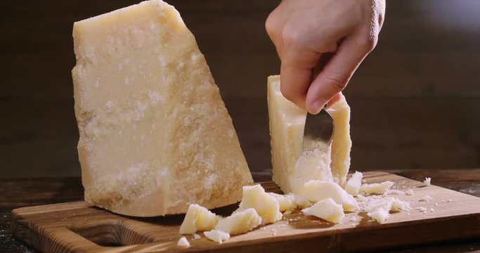 Parmesan cheese composition, on a wooden cutting board. One hand takes the knife and breaks a couple of pieces to savor the quality. Concept of: italy, cheese and tradition.