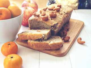 Hommade banana cake with cashew nut on top, fresh orange and apple on white wooden ground