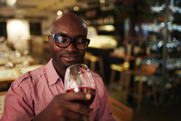 Young man in eyeglasses enjoying taste of red wine while sitting in restaurant